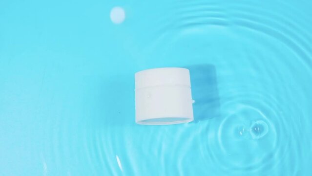 advertising slow motion, blank cosmetic cream jar on the surface of the water. Top view of drop falls into water and diverging circles of water on blue background. packaging for design
