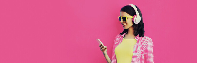 Portrait of happy smiling young woman in headphones listening to music with smartphone on pink...