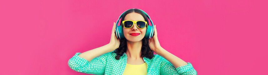 Portrait of happy smiling woman listening to music with headphones wearing sunglasses on pink background