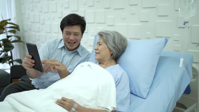 Son visiting  his mother in hospital and let her talk to family on a video call on digital tablet.