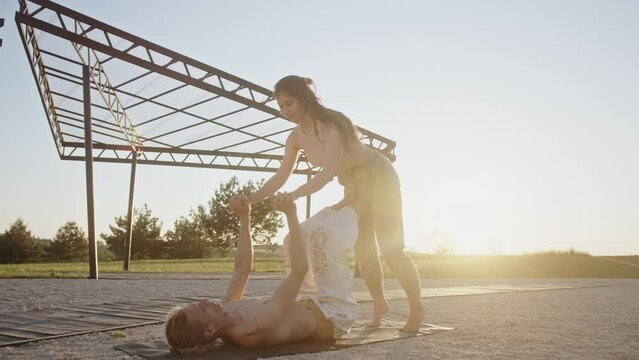 Couple doing acro yoga in park. Shirtless man lying on ground and lifting girlfriend on legs while practicing acro yoga against sunrise sky in summer morning in park