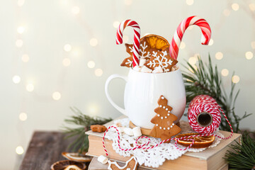 Hot winter drink in a white mug: cozy home composition with homemade gingerbread cookies, candy cane, fir tree branch. Wooden background, christmas lights and candles