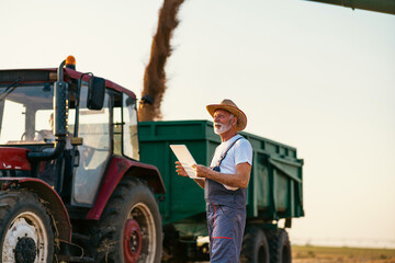 An elderly farmer with a digital tablet in his hands supervises the unloading of wheat from a...