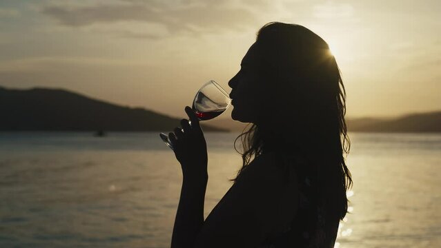 Girl drinking wine at sunset by the sea. Woman with a glass of wine by the ocean on vacation relaxes and rests.