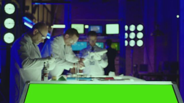 Three professional male scientists in white coats working inside modern , futuristic laboratory . In the foreground is a monitor with a green screen background . Physicians working inside blue room