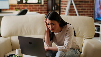 Stressed woman working remotely on laptop while trying to understand job assignment. Worried asian person looking confused at computer screen while doing remote work from home.