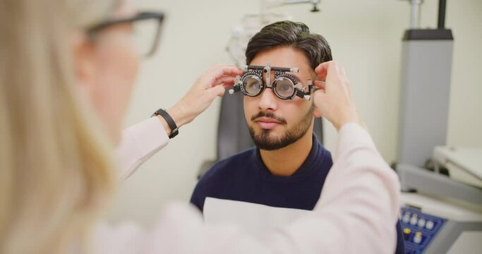 Optometrist giving male patient an eye test in consultation office. Female ophthalmologist uses spectacle frame with adjustments to check vision of a young Indian man. Optician using trail frame