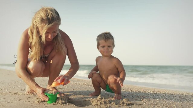 Mom with glasses plays with a little toddler sitting on the beach near the sea