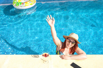 young woman with red hair smiling, waving from inside the pool, cooling off on a sunny day. young girl on summer holiday sunbathing by the pool. concept of summer and leisure time.