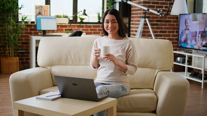 Joyful asian woman sitting in living room working from home while smiling at camera. Happy young adult student inside modern apartment having laptop on table while waiting for online class to start.