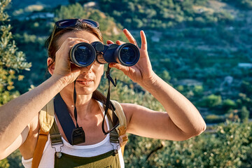 Tourist woman resting in olive grove, looking through binoculars at the mountains view. Traveling or exercising outdoors. Active people, hiking, healthy lifestyle and harmony concept.Unity with nature