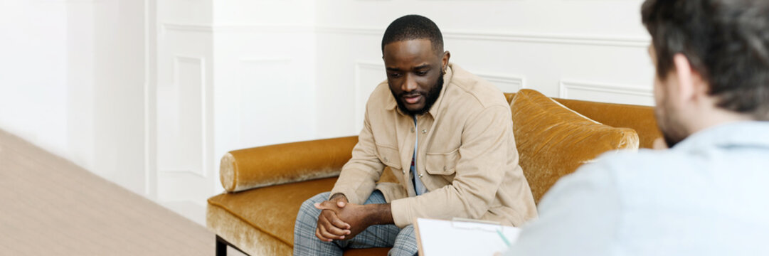 Unhappy Young Black Man Gay Having Session With Professional Psychologist At Mental Health Clinic. Professional Psychological Help Concept. Web Banner