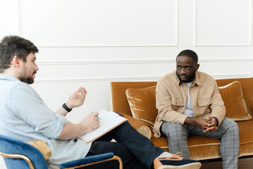A black male patient undergoing a psychotherapy session with a counselor at a mental health clinic. Young man with emotional problems consults professional therapist - obrazy, fototapety, plakaty