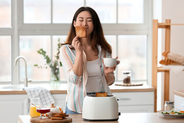 Beautiful young Asian woman with coffee and tasty toasts in kitchen - obrazy, fototapety, plakaty
