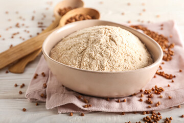 Bowl of flour with buckwheat grains and napkin on white wooden background