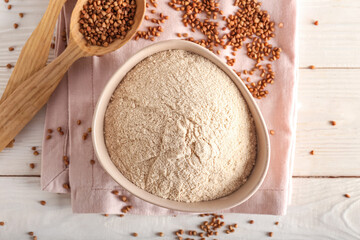 Bowl of flour with buckwheat grains and napkin on white wooden background