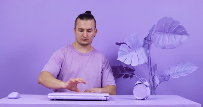 Young man adjusts to working mood by tapping fingers on table. Then he opens laptop, takes computer mouse in hand and starts working or playing video game, front view. Everything is painted purple