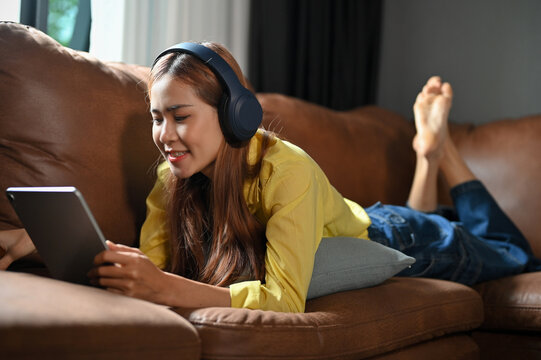 Relaxed Asian girl laying on sofa, listening music on headphones and using tablet