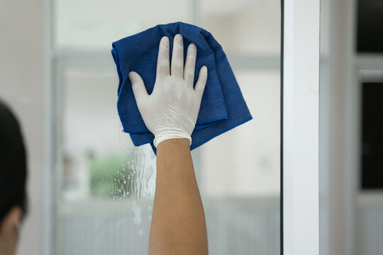 Asian woman housewife wearing rubber white gloves and holding blue cleaning clothe for wiping door glasses, cleaning up office.Woman doing housework and wipeing the glass.