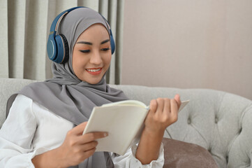 Asian Muslim woman wearing hijab and headphones reading a book in the living room.