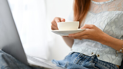 Beautiful young woman using laptop computer while holding a cup of hot coffee. cropped