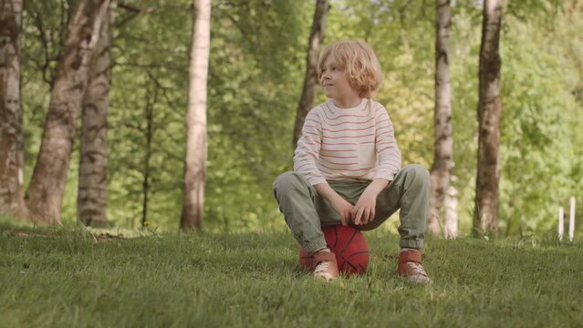 Slowmo portrait of cute blonde 7 year old boy sitting on basketball on green grass in park smiling at camera