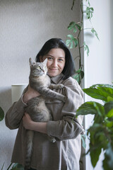 A young woman holds a gray cat with yellow eyes in her arms. The cat and her mistress at home. Taking care of pets.
