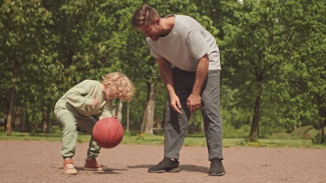 Slowmo of blonde little boy bouncing basketball off ground while playing with dad in park in summer