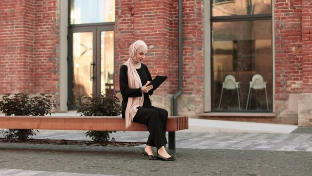Portrait of smiling happy muslim woman relaxing using digital tablet while sitting on bench. Young muslim girl looking at screen of tablet speaking during online video call on social media on city.
