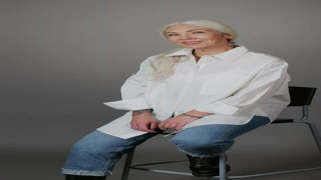 Vertical medium portrait of confident mature woman in oversized white shirt and blue jeans with arm tattoo posing for camera sitting on chair on grey studio background