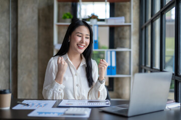 Fototapeta premium Beautiful Asian businesswoman who is excited about her laptop computer and accomplished and accomplished goals, happy and satisfied with her.