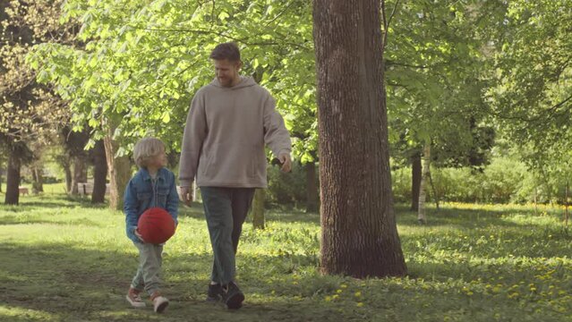 Tracking Slowmo Of Man With 7 Year Old Son Walking Along Park In Sunlight On Summer Day, Chatting And Throwing Football To Each Other