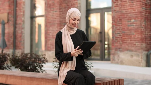 Cheerful young islamic businesswoman working on tablet while sitting near her office. Beautiful young woman with hijab working using tablet during break.