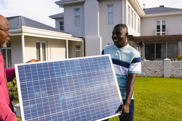African american man with senior father carrying solar panel while standing against house in yard