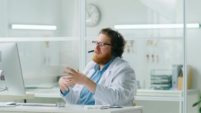 Young male doctor in headset speaking with patient via web call on computer and taking notes while working online at desk in medical office