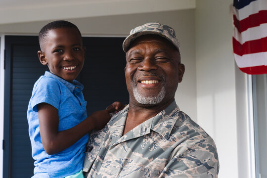 Portrait of african american smiling military senior man carrying biracial grandson against house