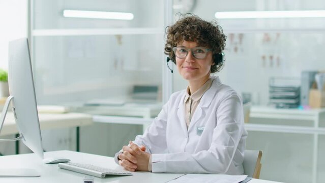 Portrait of cheerful female doctor in headset and white coat posing for camera while sitting at desk with computer in clinic