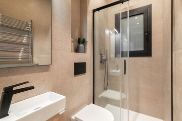 Simple bathroom with black faucet, white toilet, wooden furniture and beige tiles. Modern interior of refurbushed apartment.