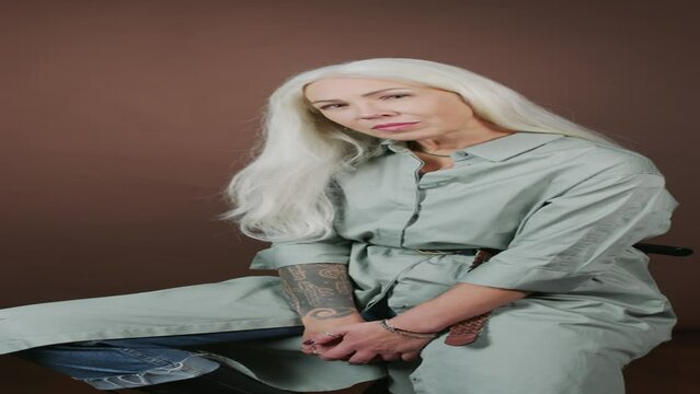 Vertical portrait of contemporary mature woman with long gray hair sitting on chair at studio background