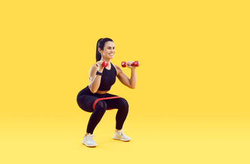 Fototapeta na wymiar Smiling active young woman athlete or coach make squats with dumbbells and fitness bands. Happy toned female trainer workout exercise on yellow studio background. Sport lifestyle.