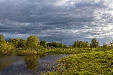 Fototapeta na wymiar Rural landscape on a spring evening. the dramatic sky is reflected in the river. Bright sun rays illuminate the grass in the foreground.