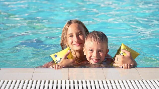 Close up of happy children swimming in pool. Brother and sister. Young beautiful girl and little boy laugh and smile broadly. Boy in inflatable armbands is holding onto side of pool. Look into camera.