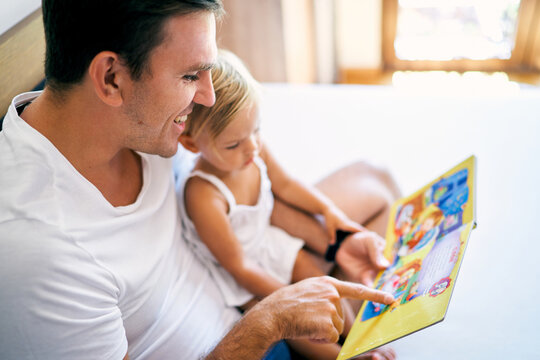 Smiling dad reads a book to a little daughter on the bed