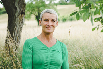 Naklejka na ściany i meble pretty, strong, powerful middle aged woman with green shirt, colorful flower pants, short grey hair posing in green meadow in front of walnut tree