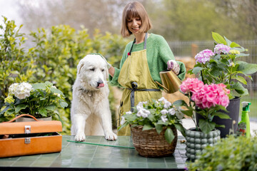 Young woman playing with her white dog while taking care of plants and flowers in the garden. Concept of happy leisure time with pets and hobby