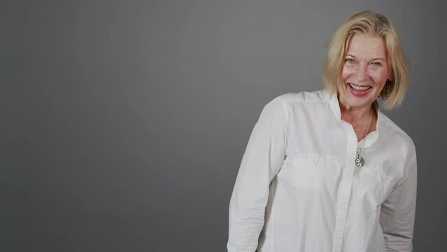 Medium slowmo portrait of attractive mature blonde woman laughing at camera standing on grey studio background