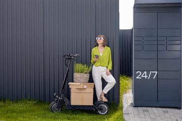 Young woman standing with phone near automatic post terminal, delivering goods by electrical...