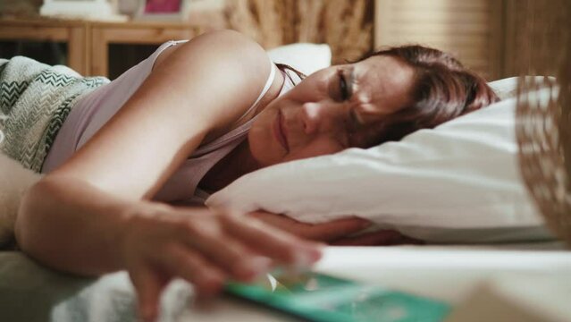 Adult woman turning off alarm on smartphone and keep sleeping. Mature sleepy woman sleeping in bed delaying alarm clock in morning