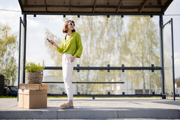 Young woman stands with parcels and flowerpot on a bus stop and using phone. Concept of...