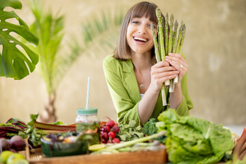 Portrait of a young cheerful woman holds a bunch of asparagus while sitting by the table full of...